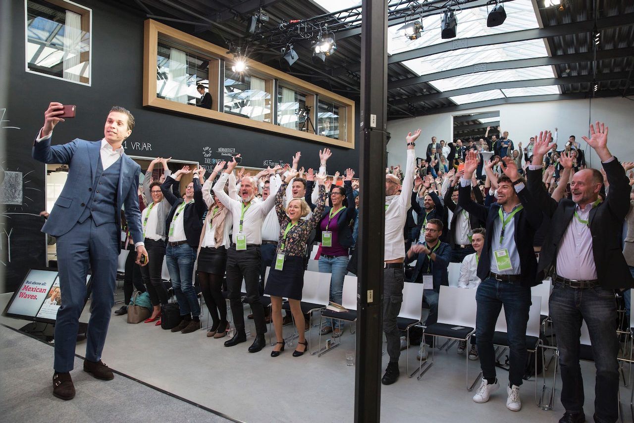 Polle on stage in front of a full room with everyone's hands in the air