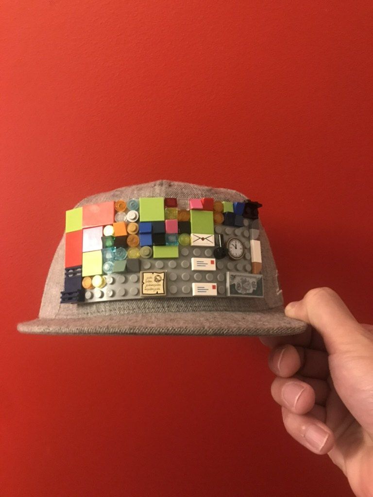 A grey cap with LEGO plates and bricks built onto the top, held up against a red wall